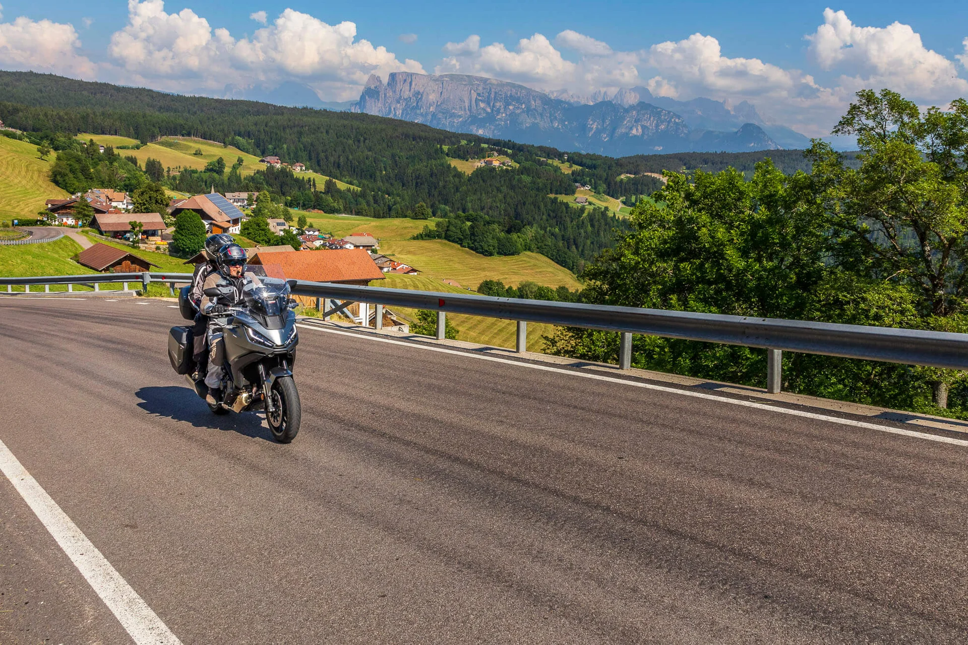 Motorradfahrer auf kurviger Bergstraße mit Dorf und Bergen im Hintergrund