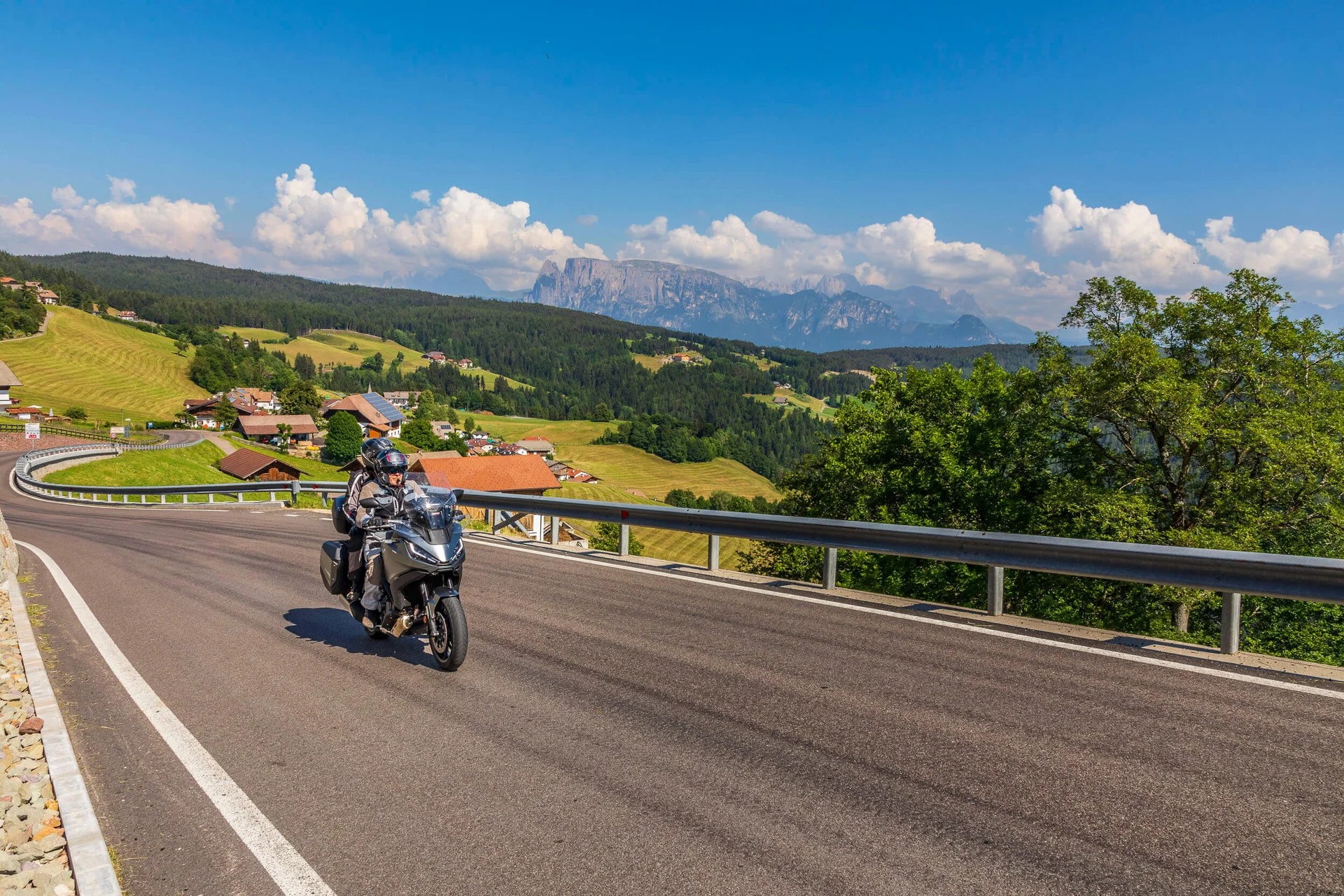 Motorradfahrer auf kurviger Bergstraße mit Dorf und Bergen im Hintergrund