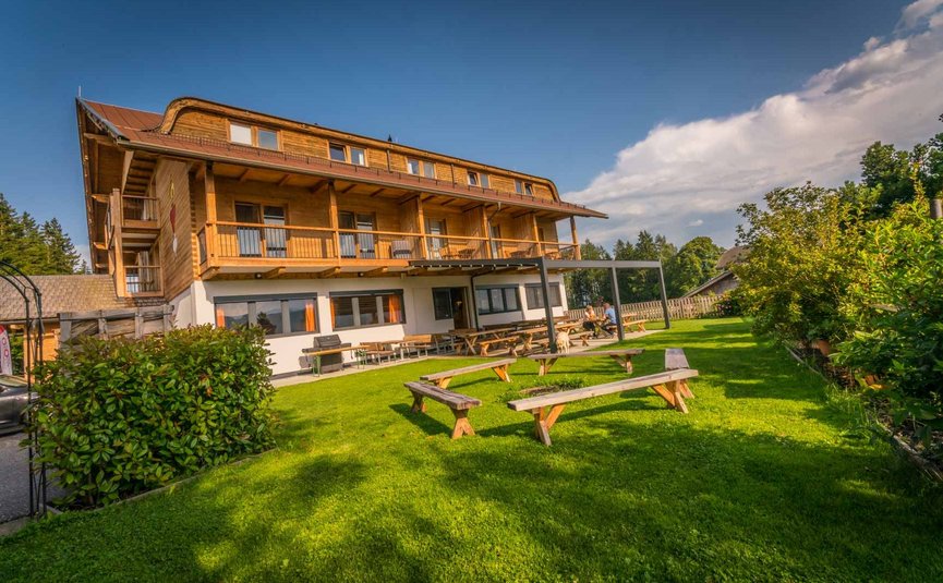 Holzhaus mit Terrasse und Sitzbänken auf grünem Rasen unter blauem Himmel