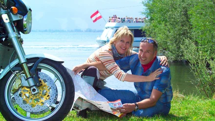 Geschichte der MoHo's Paar sitzt auf Wiese am See mit Motorrad und hält eine Karte