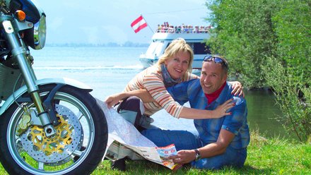 Paar sitzt auf Wiese am See mit Motorrad und hält eine Karte
