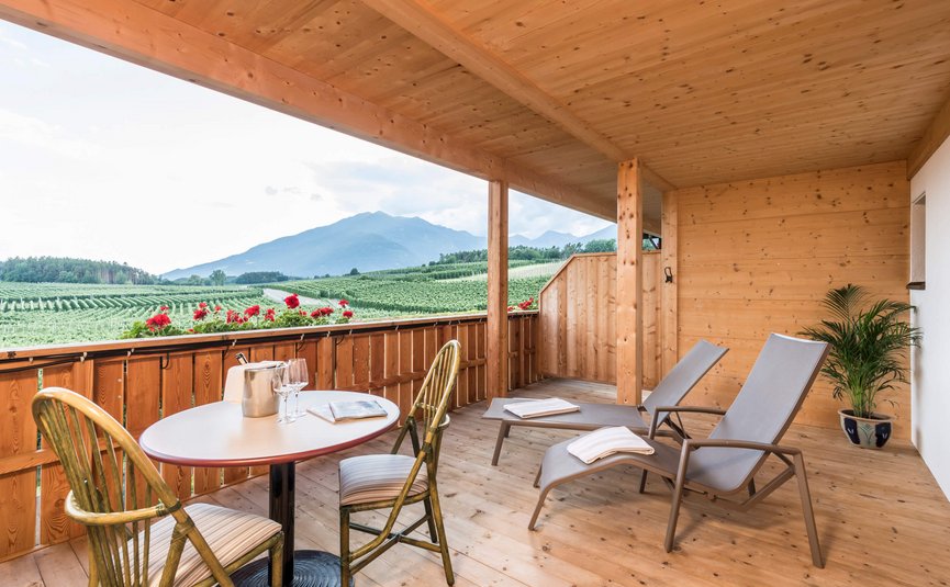 Holzbalkon mit Blick auf grüne Felder und Berge, mit Tisch, Stühlen und Liegestühlen