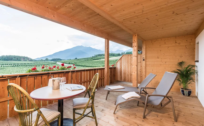 Wooden balcony overlooking green fields and mountains with table, chairs, and loungers
