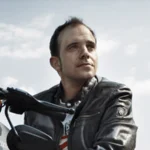 Mann in Lederjacke sitzt auf schwarzem Motorrad mit Helm daneben