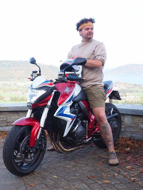 Mann lehnt an rotem Motorrad mit Bergpanorama im Hintergrund