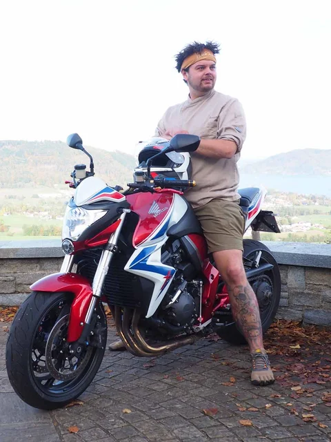 Man leaning on red motorcycle with mountains and lake in background
