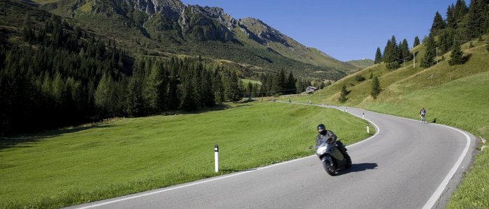 Motorradfahrer auf kurviger Bergstraße mit grünen Wiesen und Bergen im Hintergrund