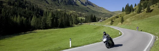 Motorradfahrer auf kurviger Bergstraße mit grünen Wiesen und Bergen im Hintergrund