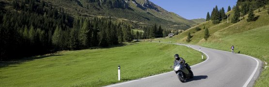 Motorradfahrer auf kurviger Bergstraße mit grünen Wiesen und Bergen im Hintergrund
