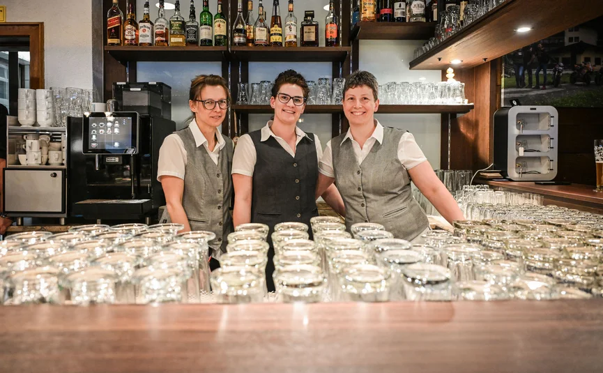 Three waitresses standing behind a bar with many glasses