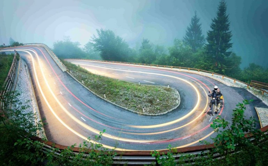 Motorrijder op kronkelende bergweg met lichtsporen in mistig landschap