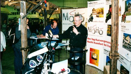 Mann präsentiert Motorrad auf Messe mit Informationsständen im Hintergrund