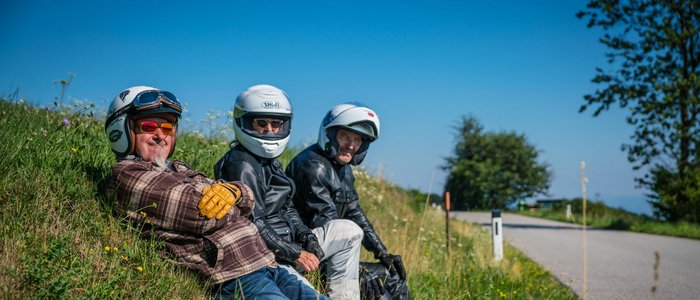 Drei Motorradfahrer in Helmen sitzen auf einer Wiese neben einer Straße.