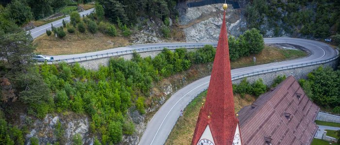 Kirchturm mit rotem Dach neben kurviger Bergstraße und Wald