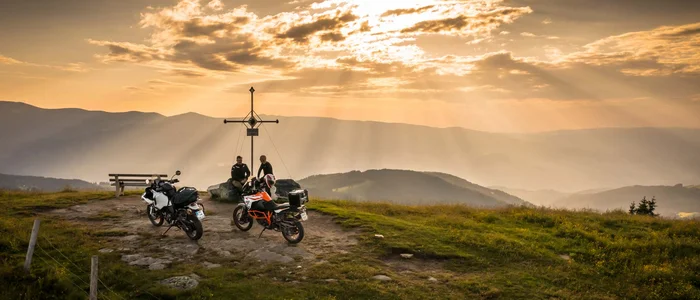 Two motorcycles and two people by a summit cross at sunset in the mountains