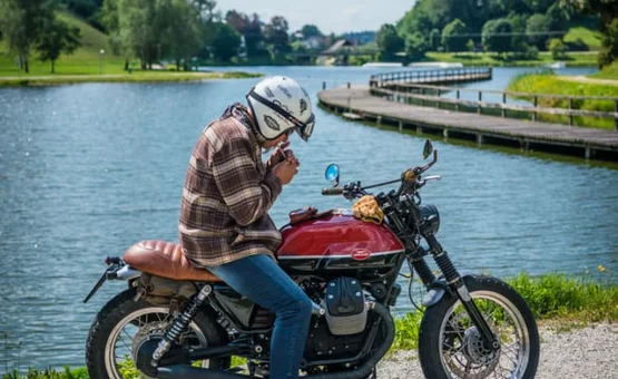 Mann mit Helm sitzt auf Motorrad am See und zündet eine Zigarette an