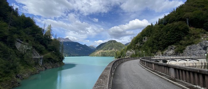 Straße am Staudamm neben blauer Bergsee und bewaldeten Bergen