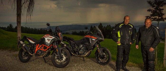 Zwei Männer in Motorradkleidung neben zwei KTM-Motorrädern vor dunklen Wolken