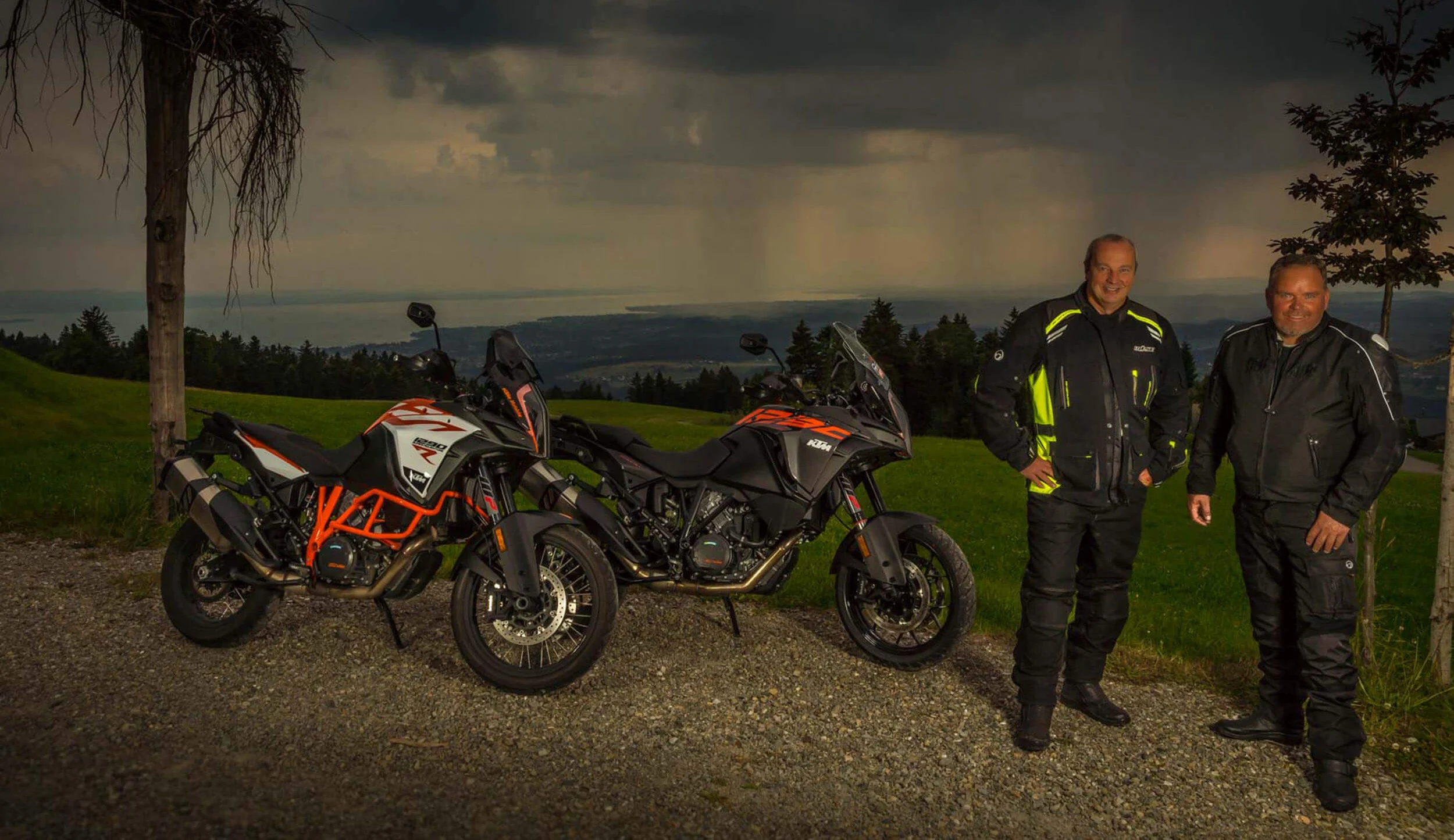 Two men in motorcycle gear standing next to two KTM motorcycles under dark clouds