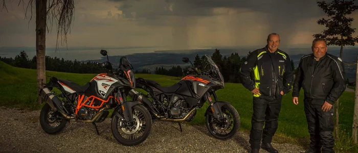 Two men in motorcycle gear standing next to two KTM motorcycles under dark clouds