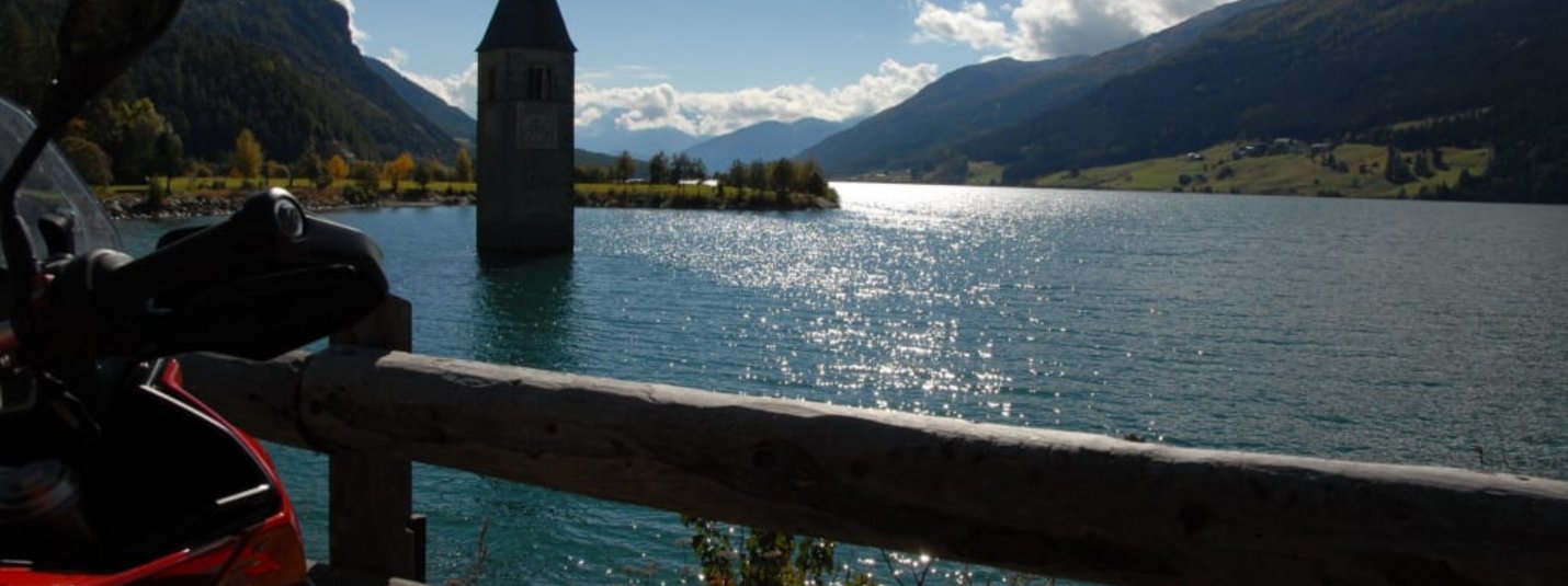 Kirchturm ragt aus einem See in den Alpen mit Motorrad im Vordergrund