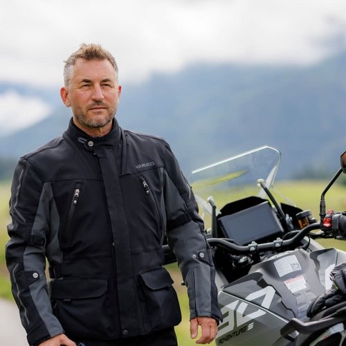 Mann in schwarzer Motorradkleidung hält Helm neben Motorrad auf Landstraße