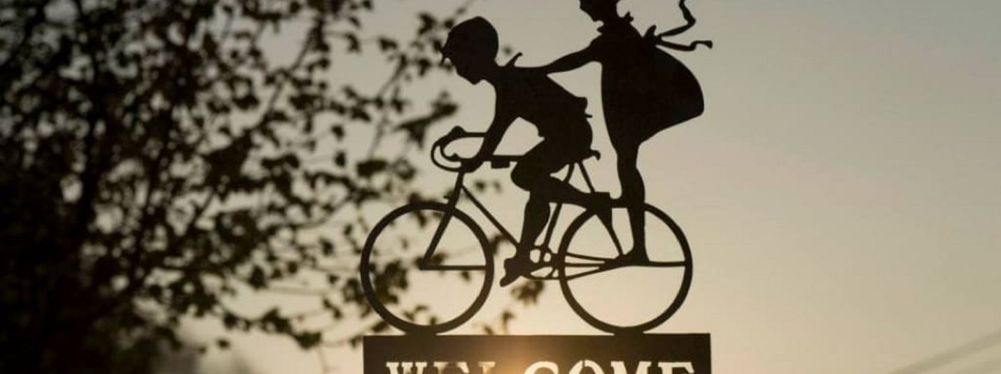 Metallfigur von zwei Personen auf Fahrrad über Willkommensschild bei Sonnenuntergang