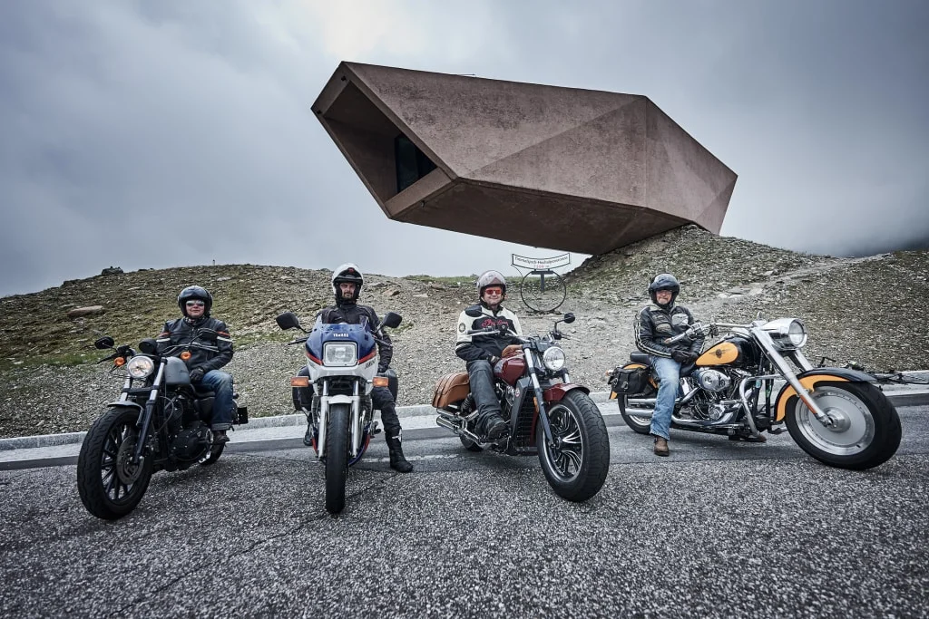 Four motorcyclists in front of futuristic building on mountain road under cloudy sky