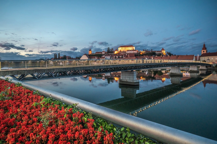 Carinthia–Styria Wine Country Ride Beleuchtete Brücke und Schloss mit Stadt am Flussufer bei Dämmerung