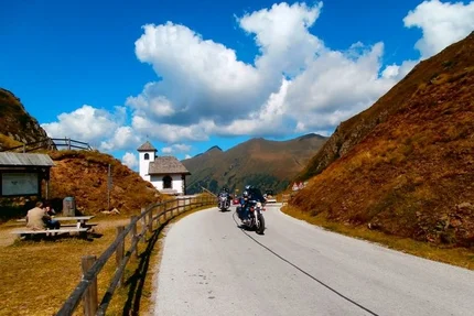 Motorcyclists riding on mountain road beside chapel under sunny sky