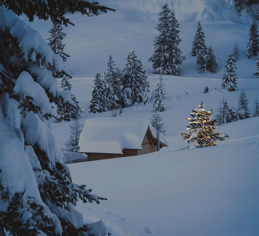 Schneebedeckte Hütte und beleuchteter Tannenbaum in verschneiter Winterlandschaft