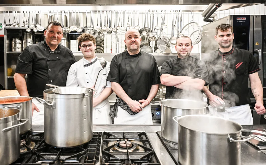 Five chefs standing in a professional kitchen with steaming pots.
