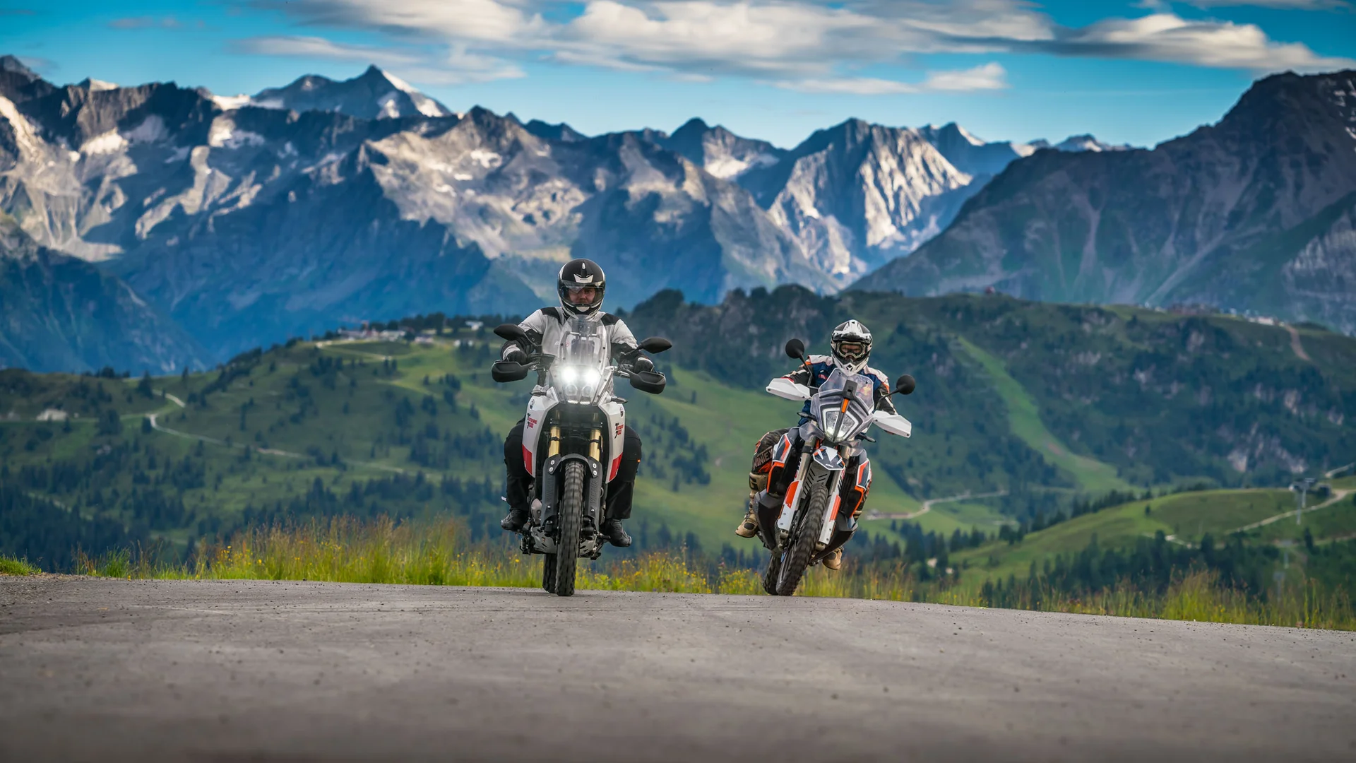 Two motorcyclists riding on mountain road with Alps in background