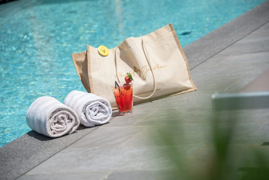 Two white towels, a cocktail, and a bag beside a pool