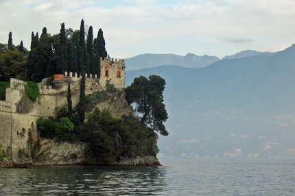 Old castle on lakeshore with cypress trees and mountains in background