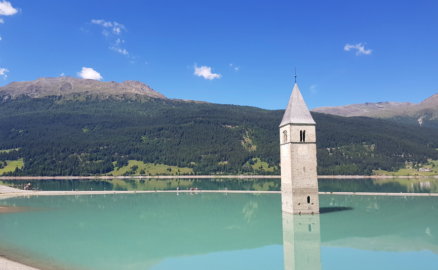 MoHo Schönauer Hof- Tour 12 Stilfserjoch Umbrail Church tower emerging from a lake with mountains and blue sky in the background
