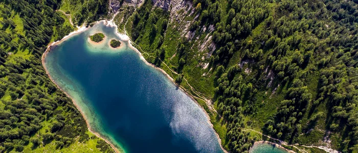 Luftaufnahme eines Bergsees mit zwei kleinen bewaldeten Inseln und umliegendem Wald