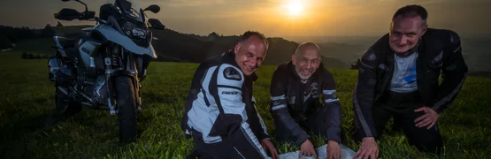 Drei Motorradfahrer schauen auf eine Landkarte bei Sonnenuntergang auf einer Wiese