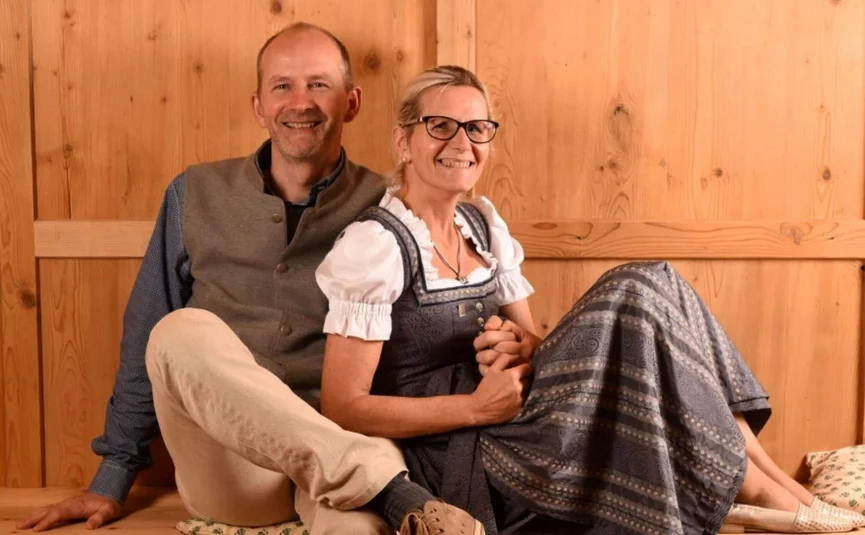Smiling couple in traditional clothing sitting on wooden bench against wooden wall
