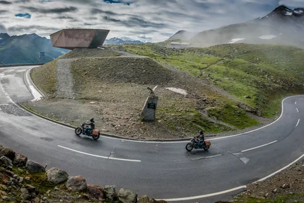 Kurvenreiche Bergstraße mit zwei Motorradfahrern und Aussichtspunkt im Nebel