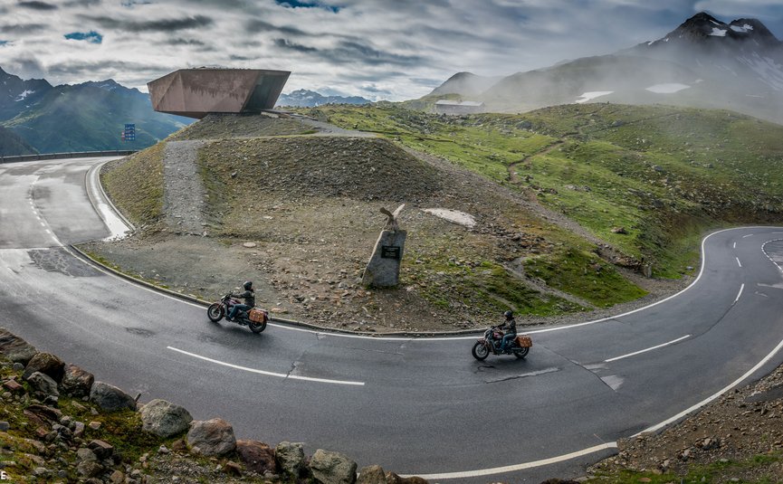 Kurvenreiche Bergstraße mit zwei Motorradfahrern und Aussichtspunkt im Nebel