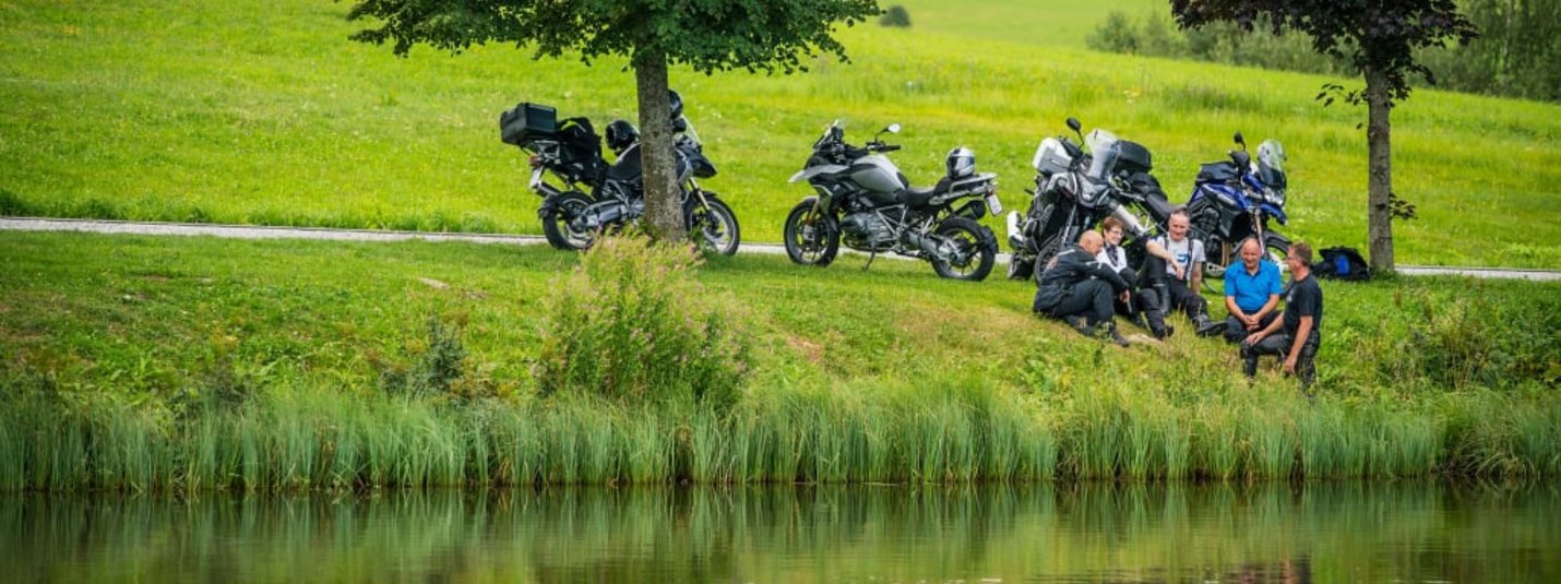 MOTORRAD - Genuss-Tage So-Do 2026 Fünf Menschen sitzen am Seeufer neben Motorrädern auf einer grünen Wiese