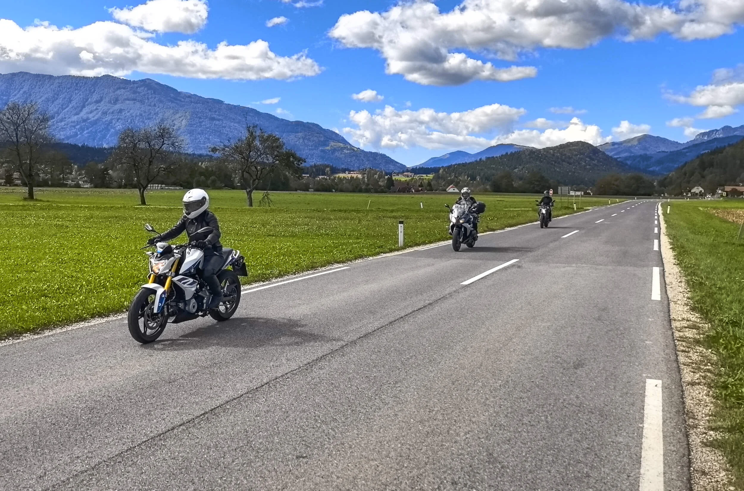 Drie motorfietsers rijden op een landweg met bergen op de achtergrond