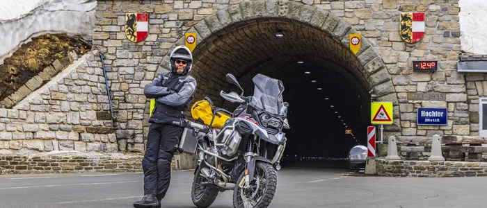 Motorcyclist standing beside bike in front of stone tunnel entrance with snow