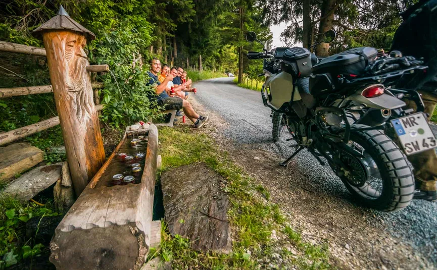Group of people sitting by forest roadside near motorcycle and wooden water trough