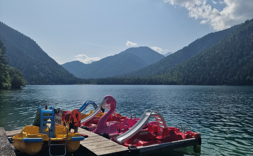 Tretboote mit Rutschen am See vor bewaldeten Bergen unter blauem Himmel