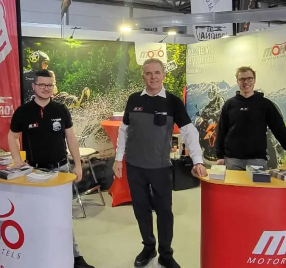 Three men at trade show booth with motorcycle info and brochures