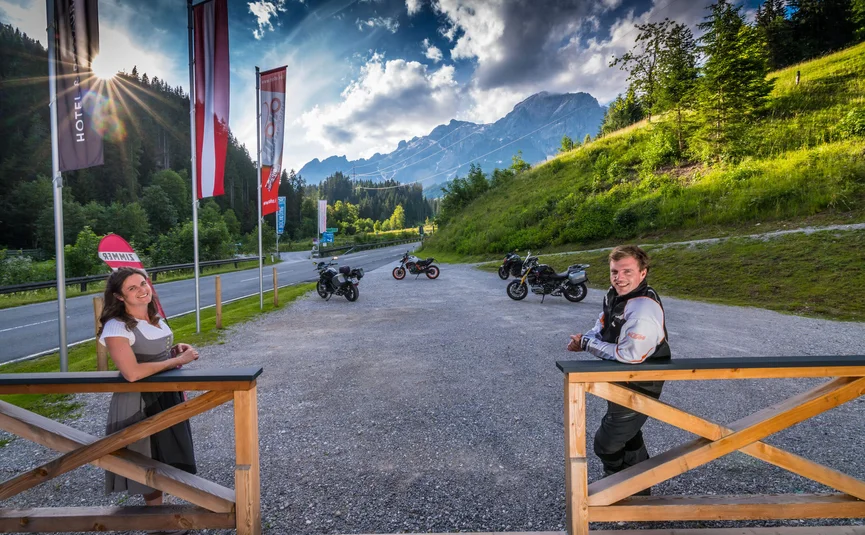 Man and woman by wooden gate with motorcycles and mountains on sunny day