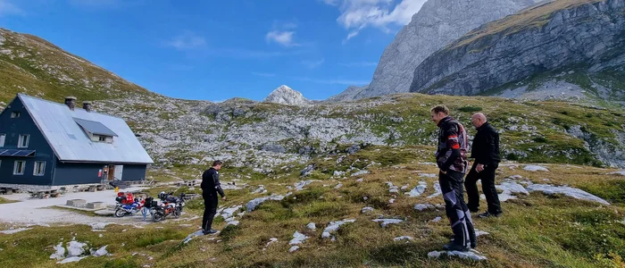 Three men in motorcycle gear hiking in mountainous landscape near cabin