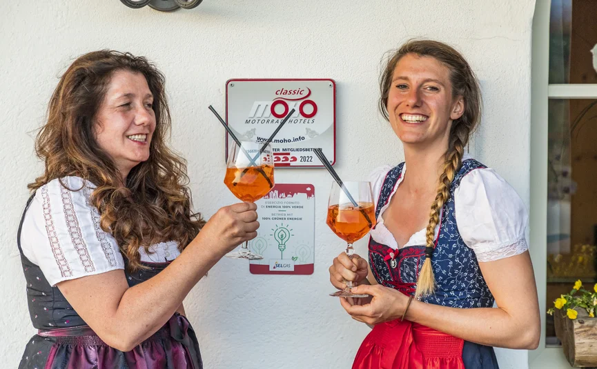 Two women in dirndls toasting with Aperol Spritz in front of white wall